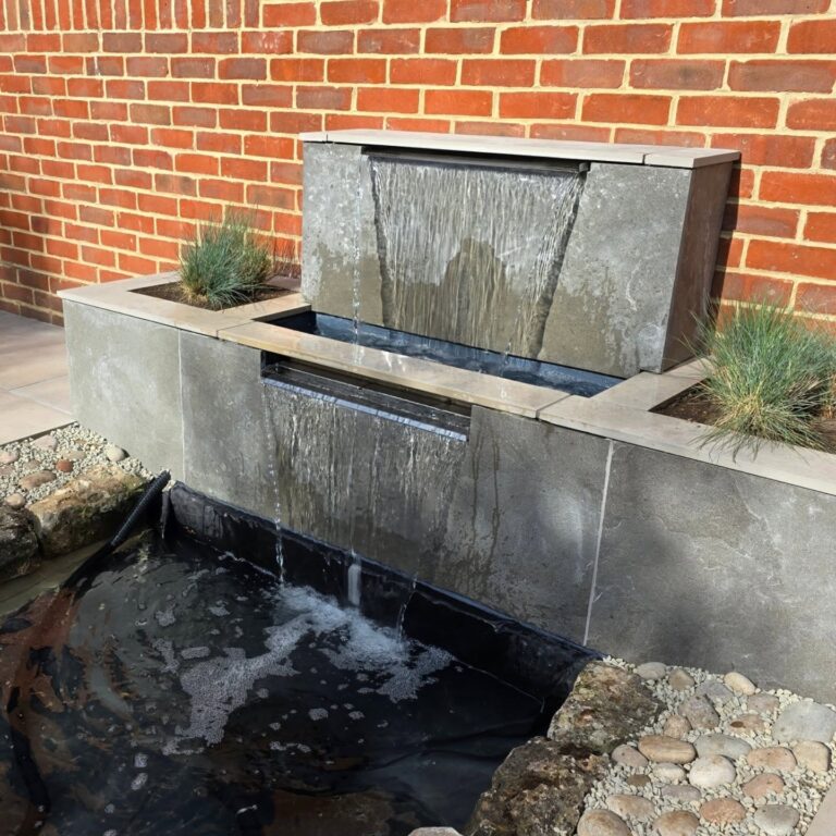 Modern water feature with cascading water flowing from a porcelain-clad unit into a lower pond, set against a brick wall with planted grasses