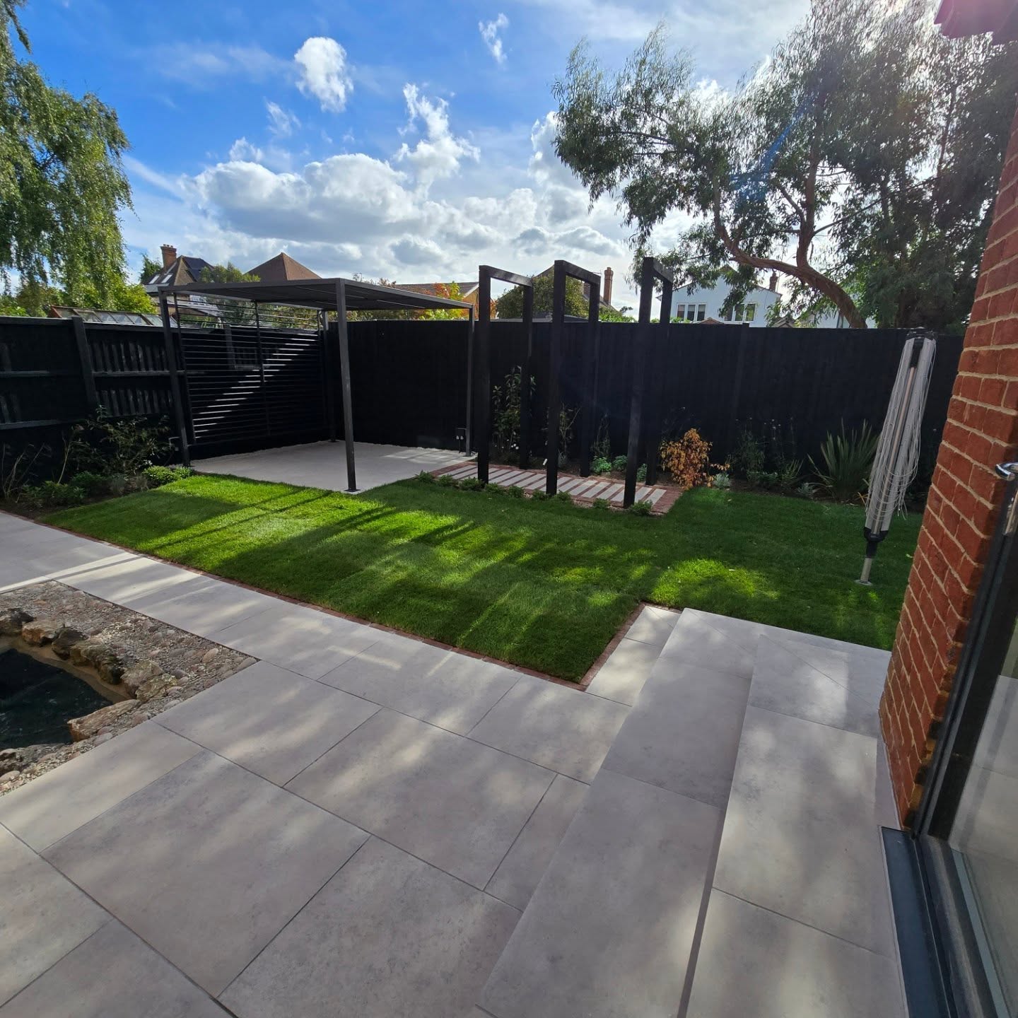 Overview of contemporary garden with porcelain paving, lush lawn, and black pergola frames